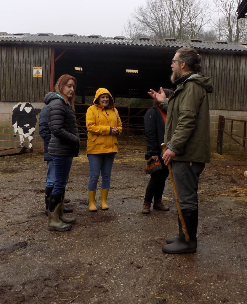 A group of people standing in a farmyard, one of them is talking to the rest