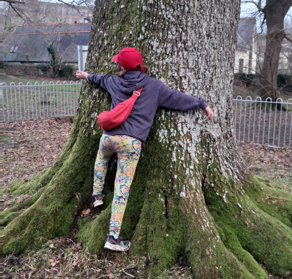 A young person wearing a red hat and bag with their arms around the trunk of a tree.