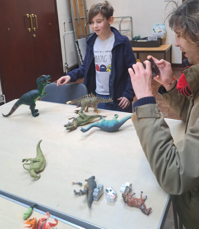 A boy standing at a table with toy dinosaurs. A man is taking a picture.
