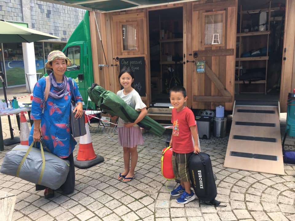 A woman and two children standing outside a mobile shed, holding camping equipment.