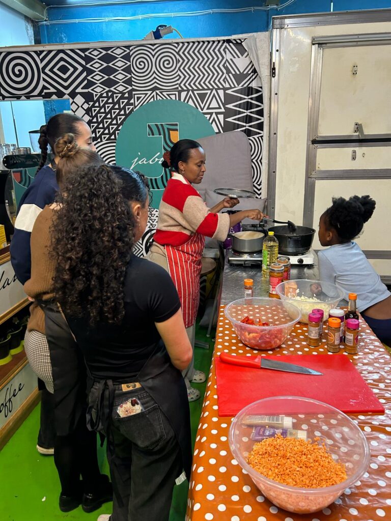 A Black woman cooking at a hob while other women and girls watch.